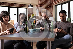 Multiracial friends sitting at table in living room using on smartphones while surfing internet.