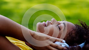 Multiracial girl lying down in grass and enjoying music in headphones, side view.