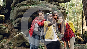 Multiracial group of friends tourists are taking selfie in forest with rocks in background using smartphone, men and