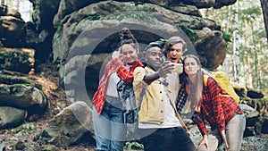 Multiracial group of friends tourists are taking selfie in forest with rocks in background using smartphone, men and