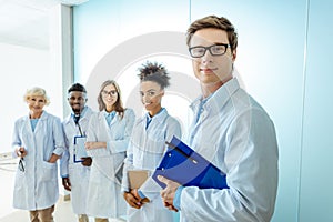 Multiracial group of smiling medical interns in lab coats standing in a row with clipboards