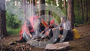 Multiracial group of young male and female travelers is cooking food roasting marshmallow on fire sitting in forest and