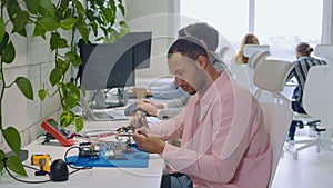 Multiracial Handsome Guy Engineer Resolders Parts of Motherboard in Office, Repairs Circuits, Checking the Chip or Board