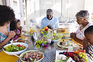 Multiracial multigeneration family with meal on table praying before having lunch at dining table