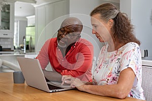 Multiracial senior couple using laptop while sitting at table in living room