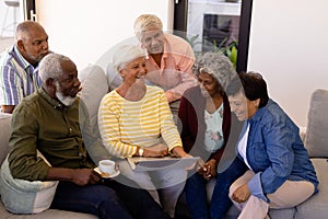 Multiracial senior woman holding laptop laughing while sitting with friends on sofa in nursing home