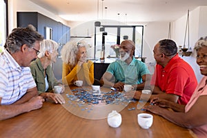 Multiracial seniors friends playing jigsaw puzzle while sitting at wooden table in nursing home