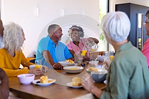 Multiracial seniors looking at happy woman serving juice to man at dining table in retirement home