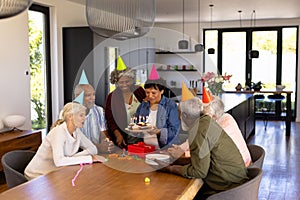 Multiracial seniors wearing party hats looking at woman holding birthday cupcakes at table