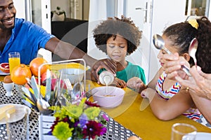 Multiracial smiling father pouring milk in breakfast cereals for children at dining table at home