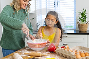 Multiracial smiling mother with daughter mixing batter in bowl with wire whisks on table in kitchen