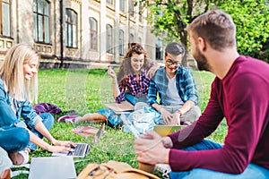 multiracial smiling students with digital devices sitting on green grass