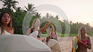 Multiracial surfer girls holding surfboards talking cheerfully standing on beach at sunrise. Happy female surfers