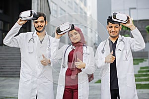 Multiracial team of doctors with virtual reality glasses, posing with thumbs up outside the hospital