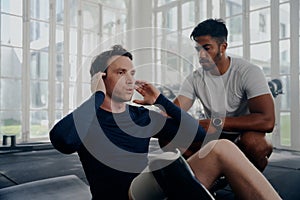 Multiracial trainer next to young man in sportswear with artificial limb doing push-ups at the gym