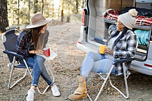 Multiracial women friends having fun camping with camper van while drinking coffee outdoor - Focus on senior african woman face