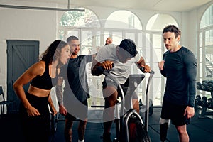 Multiracial young adults in sports clothing cheering on friend cycling on exercise bike at the gym