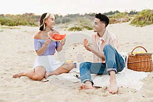 Multiracial young couple eating watermelon during picnic on beach