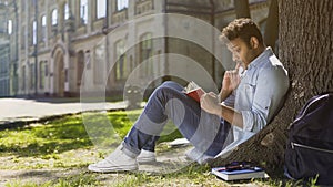 Multiracial young guy sitting under tree, reading interesting book, bookworm