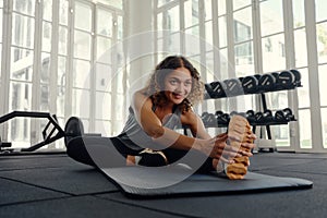Multiracial young woman in sports clothing sitting and stretching on exercise mat at gym