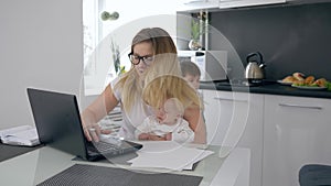 Multitasking mother with baby in arms work at table on background of jumping son at kitchen