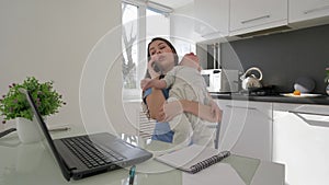 Multitasking mother with crying baby boy while working on laptop computer and talking on smartphone sitting at table in