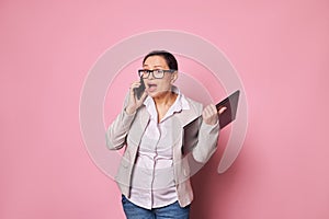 Multitasking pregnant business woman isolated over pink background, holding laptop computer talking by mobile phone. photo