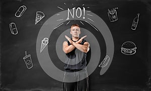 Muscled young man showing stop sign with his hands and says no to unhealthy food on the background of blackboard