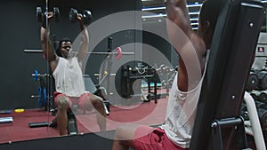 Muscular African American Man sits on bench and lifts up dumbbells in the gym.