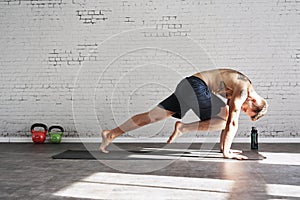 Muscular athlete exercising yoga asana in sunny sport club, brick wall. Fit shirtless male fitness model.