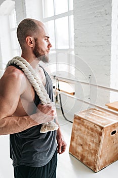 Muscular athlete holding battle ropes on shoulders in cross hall. Vertical