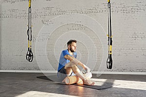 Muscular athlete sitting on mat resting after workout in sunny sport club, brick wall. Fit shirtless male fitness model.