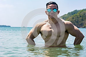 A muscular and athletic asian man wearing shades takes a dip in the sea. Summertime body goals for the beach