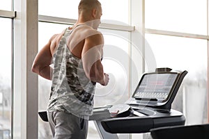 Muscular athletic bodybuilder fitness model running treadmill gym near big window