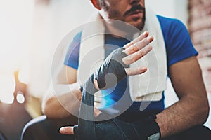 Muscular Boxer man prepairing hands for hard kickboxing training session in gym.Bearded Young athlete tying black boxing