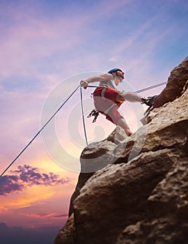 Muscular climber man in protective helmet abseiling from cliff rock wall using rope Belay device and climbing harness on evening photo