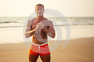 Muscular and fit young bodybuilder man doing workout warm up stretching on a paradise beach at sunset.fitness male model