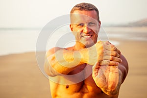 Muscular and fit young bodybuilder man doing workout warm up stretching on a paradise beach at sunset.fitness male model
