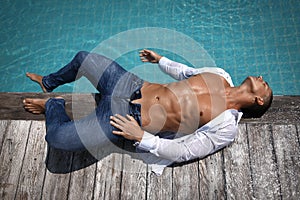 Muscular gorgeous sexy young wet man with athletic body is lies next to the pool on a wooden platform in blue jeans and white