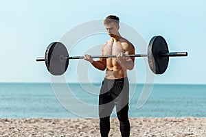Muscular guy bodybuilder doing exercises with big dumbbells outdoors on the beach, on a summer sunny day, training on the sea