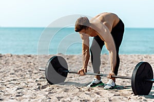 Muscular guy bodybuilder doing exercises with big dumbbells outdoors on the beach, on a summer sunny day, training on the sea