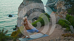 Muscular guy training yoga asana stretching at beautiful ocean cliff edge alone.