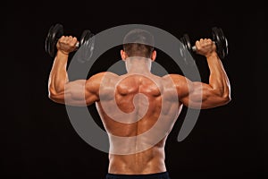 Muscular male model bodybuilder doing exercises with dumbbells, turned back. Isolated on black background with copyspace