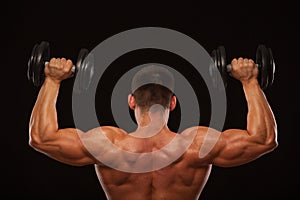 Muscular male model bodybuilder doing exercises with dumbbells, turned back. Isolated on black background with copyspace