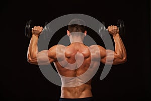 Muscular male model bodybuilder doing exercises with dumbbells, turned back. Isolated on black background with copyspace