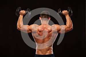 Muscular male model bodybuilder doing exercises with dumbbells, turned back. Isolated on black background with copyspace