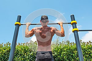 Muscular man in a cap doing pull-ups on the horizontal bar in the park outdoors, back view of his back