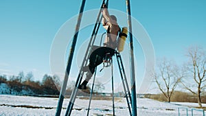 A muscular man with a drager on his back climbs on his hands up an inclined ladder on a sports field in a winter park