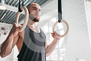 Muscular man holding gymnastic rings at light gym and looking away
