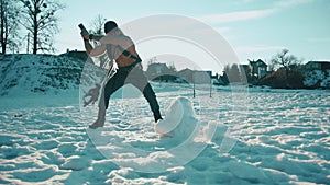 A muscular man with a naked torso and with ballon compressed air drager breaks a block of ice with a sledgehammer in the